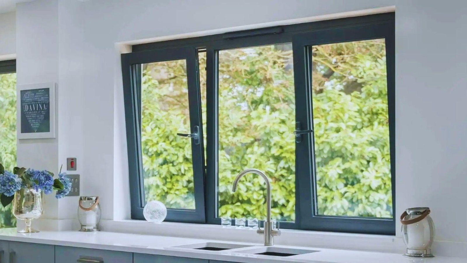 Modern kitchen with three dark gray tilt and turn windows over sink, blue hydrangeas, green trees visible outside