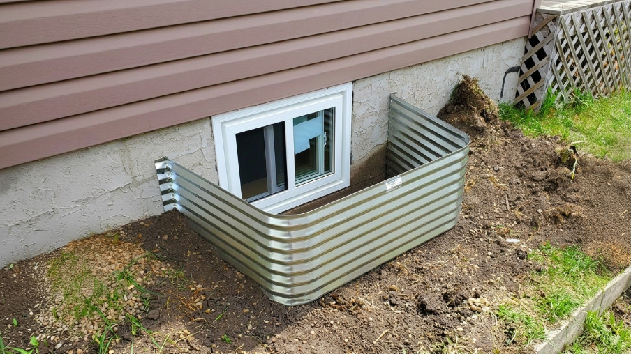 Galvanized metal window well surrounding white sliding basement egress window against stucco foundation wall with disturbed soil.