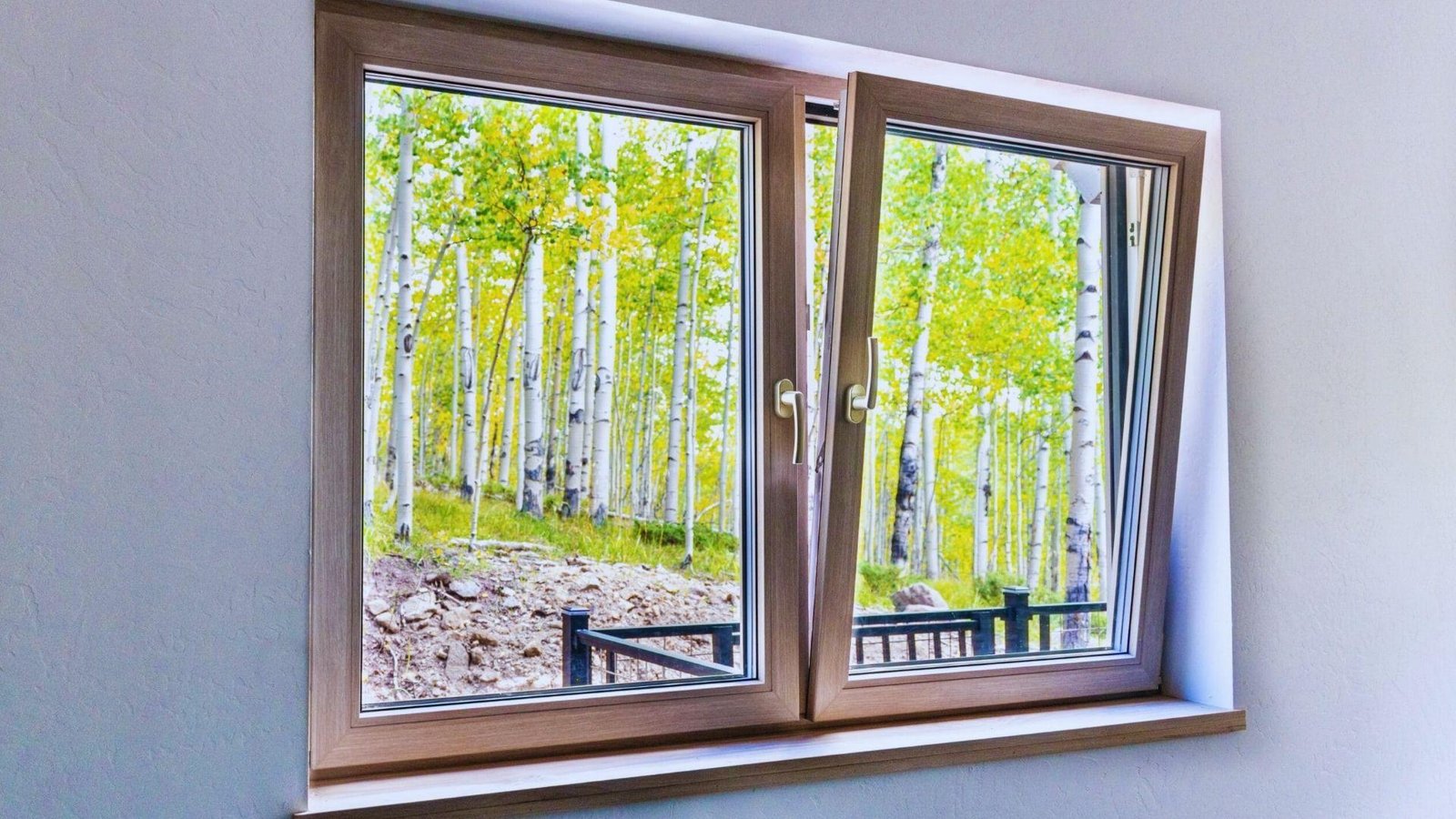 Wood-finish tilt and turn window tilted inward overlooking aspen grove with white bark, green leaves, rocky hillside