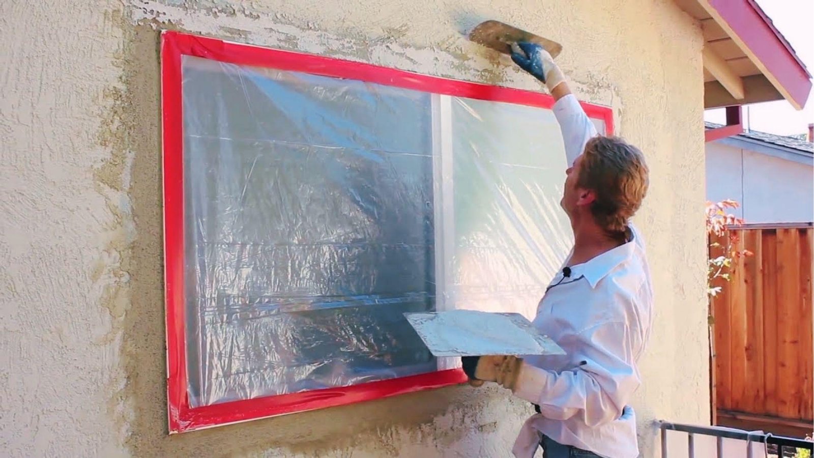 Contractor trowels fresh stucco around a masked window with red tape for smooth trim.