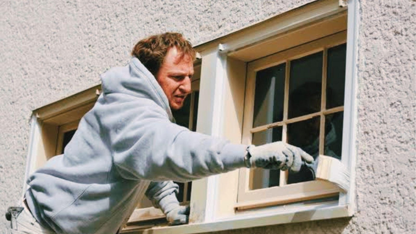 Painter brushes white trim on wood window set into rough textured stucco exterior.