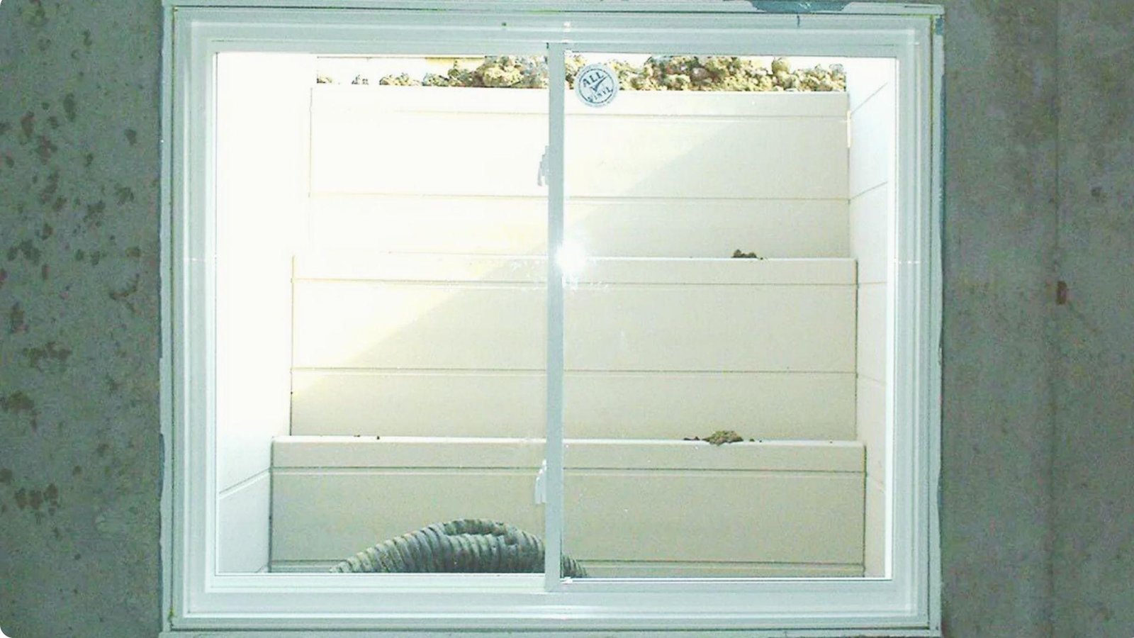 Interior view of white sliding basement window overlooking a bright plastic step window well with drainage hose visible below.