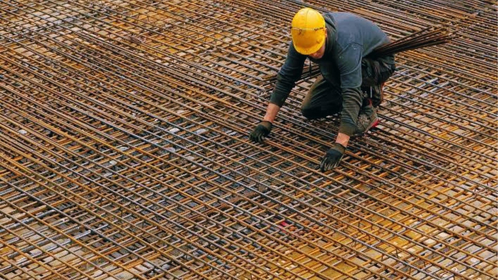 Construction worker in yellow helmet placing steel rebar grid for concrete slab reinforcement on site