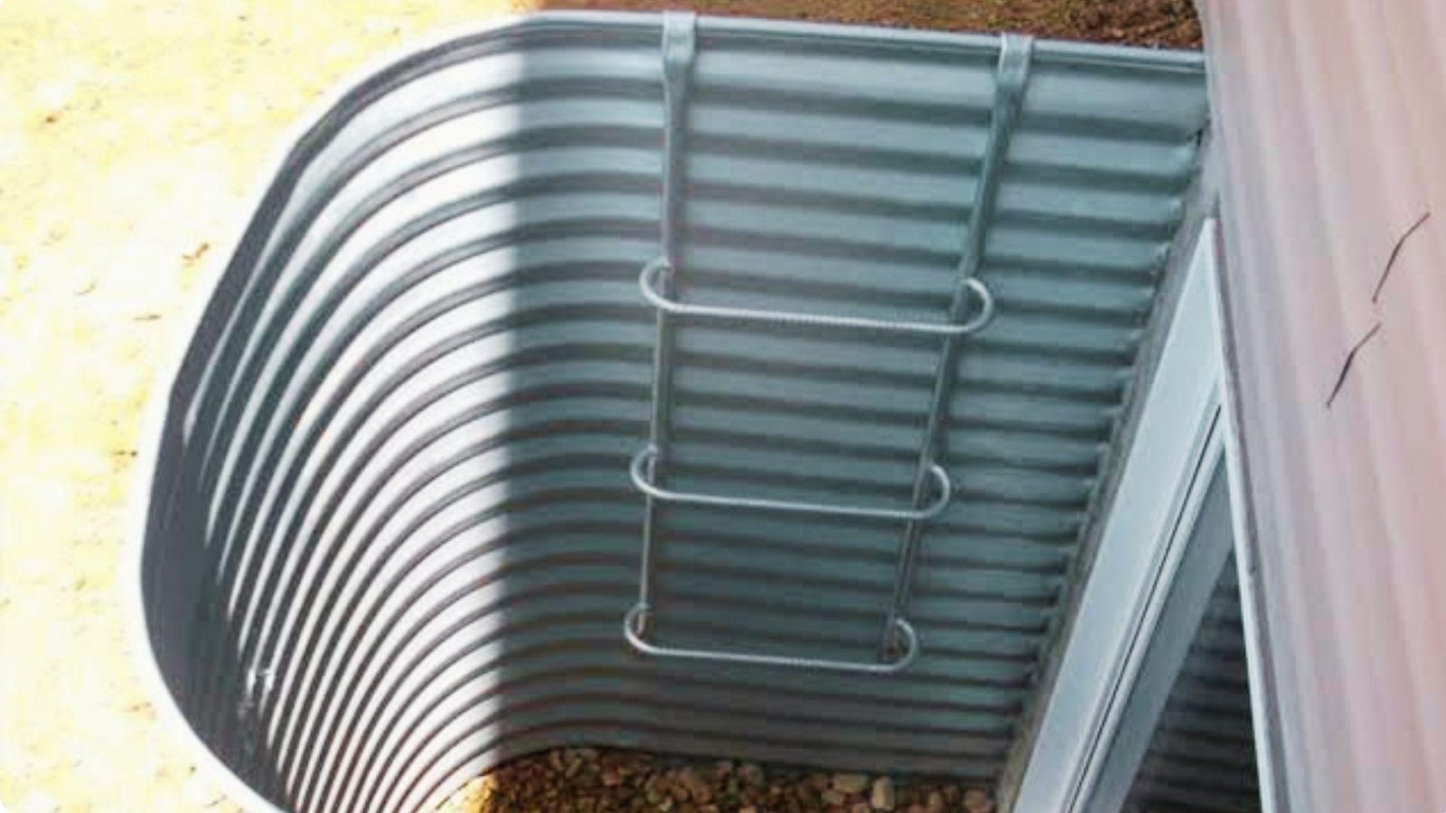 Overhead view of galvanized corrugated metal egress window well with metal escape ladder mounted on interior wall and gravel base.