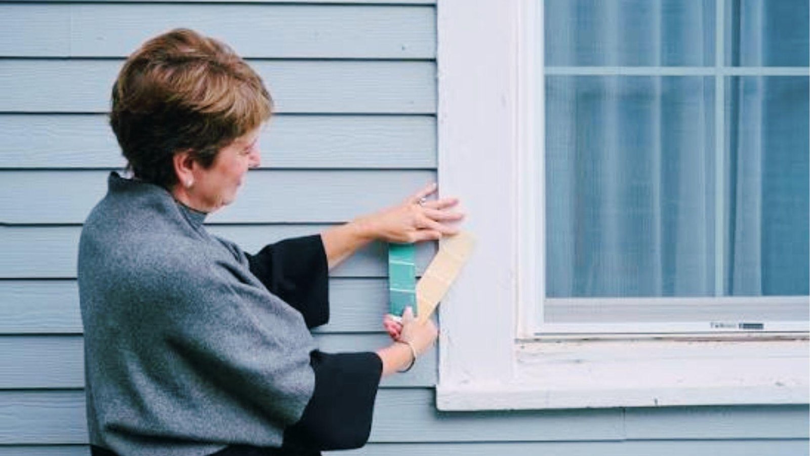 The woman compares paint swatches on white window trim against the blue siding exterior.